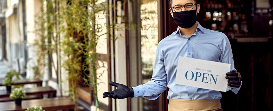 Happy waiter with protective face mask holding open sign while standing at cafe doorway.
